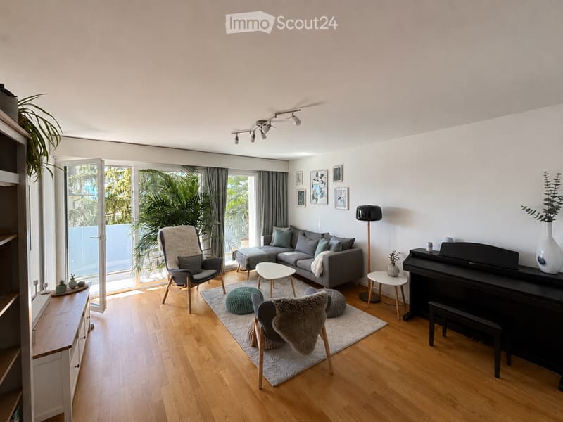 Modern living room with light wooden flooring, large windows, grey sofa, coffee table, chairs, piano, and decorative items.