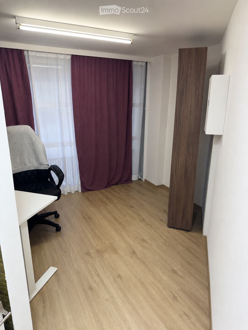 A small, furnished room with light wooden flooring, white walls, and large windows covered by dark curtains. A black office chair and a white desk are visible.