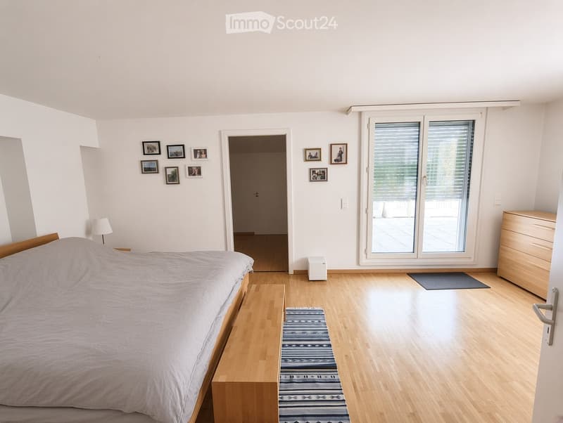 Bright bedroom with wooden flooring, large sliding glass doors leading to a balcony, a neatly made bed, a nightstand with a lamp, a dresser, and several picture frames on the walls.