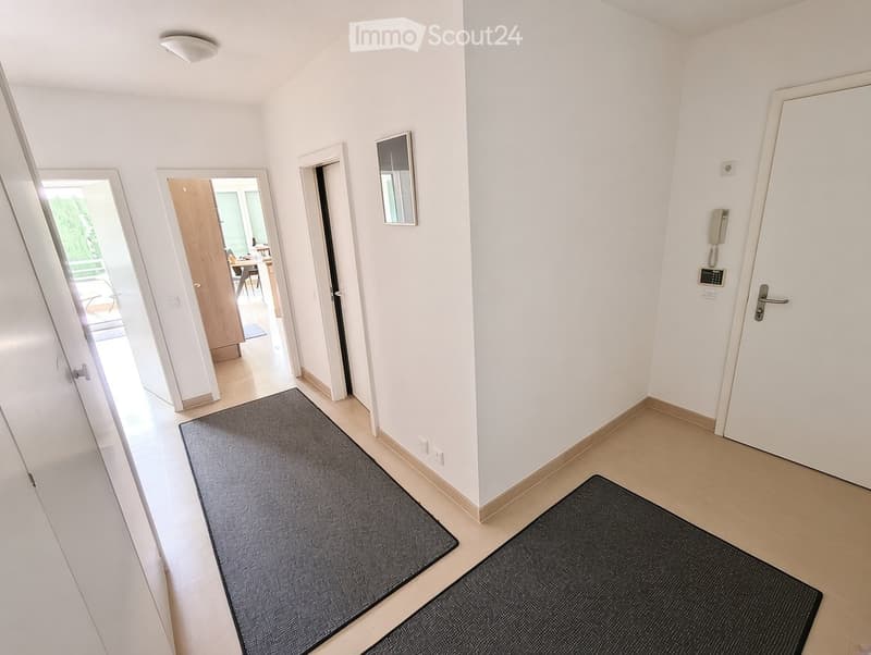 Bright hallway with white walls, tiled floor, and two black doormats. Doors lead to adjacent rooms, and a small mirror is mounted on the wall.