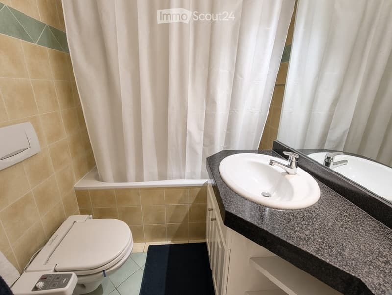 Modern bathroom with a white sink, mirror, toilet, and a dark countertop. Beige tiles and a white shower curtain are visible.
