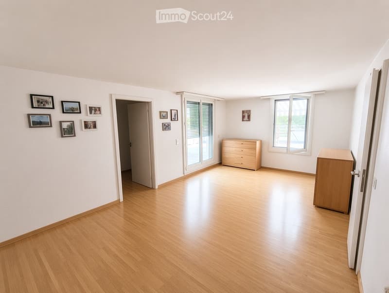 Empty room with light wooden flooring, white walls, and large windows with blinds. There are several framed pictures on the walls and a sliding glass door leading to a balcony area.
