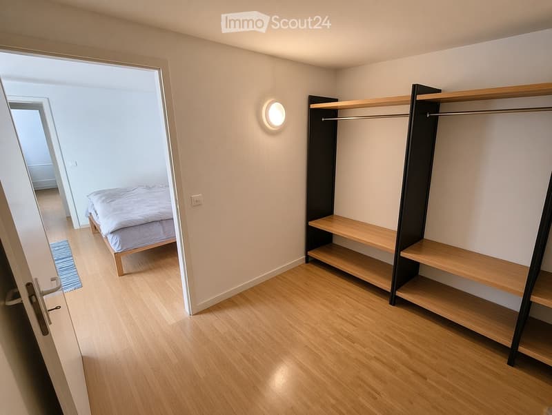 Minimalist bedroom with light wooden flooring, a single bed with white bedding, a built-in wardrobe with wooden shelves, and a round wall-mounted light fixture.