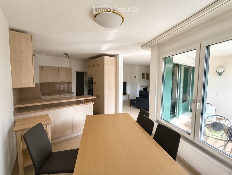 Modern, well-lit room with a dining table, chairs, a sofa, and a kitchen area with cabinets and an oven. Large windows and a balcony door provide natural light and access to the outdoor area.