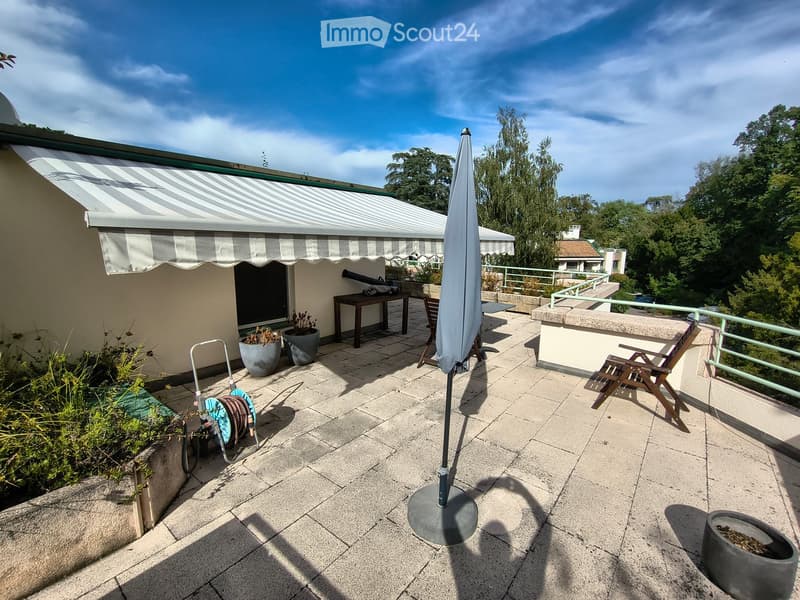 Balcony with tiled floor, covered awning, outdoor furniture including a table, chairs, and an umbrella, with potted plants and a view of greenery.