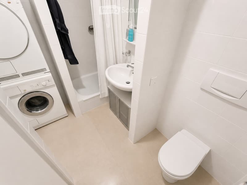 Modern bathroom with white tiles, a washing machine, bathtub, sink, toilet, and a small shelf with toiletries.