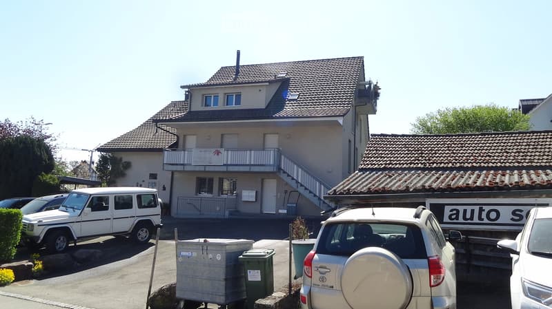 Building. Two-story residential building with a tiled roof, balcony, and several parked cars in front.