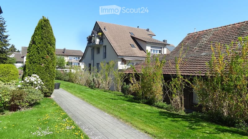 Garden / Outdoor Area. A well-maintained garden area with a paved pathway leading to a house, surrounded by green grass, shrubs, and trees. The house features a tiled roof and multiple windows, with a small balcony visible on the upper floor.