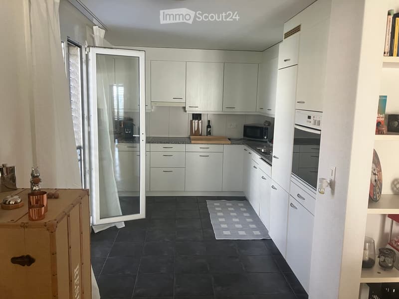 Kitchen. Modern kitchen with white cabinets, black tiled floor, and various appliances including a refrigerator, microwave, and oven. A small rug is placed on the floor, and a wooden table with copper cups is visible.