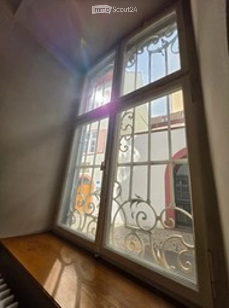 A small, unfurnished room with a large window featuring decorative wrought-iron bars. The window is framed by wooden sills and the room has a simple, clean look with white walls and a wooden floor.