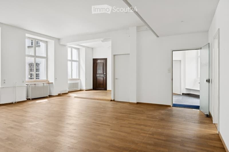 Empty room with wooden flooring, white walls, large windows, and a wooden door leading to another space.