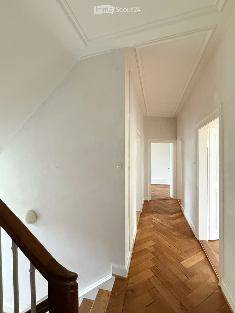 A narrow hallway with wooden herringbone flooring, white walls, and a staircase leading upwards. The hallway has several open doorways leading to other rooms.