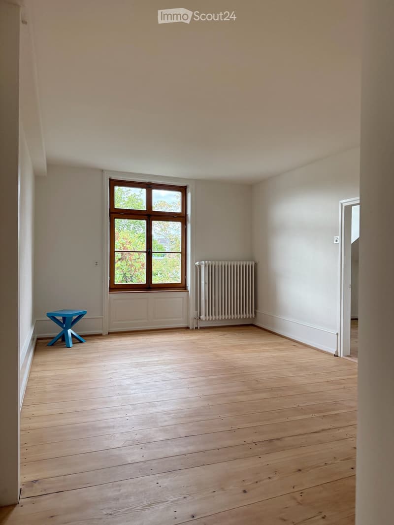 Empty room with wooden flooring, light-colored walls, a window with wooden frame, radiator, and a small blue stool.