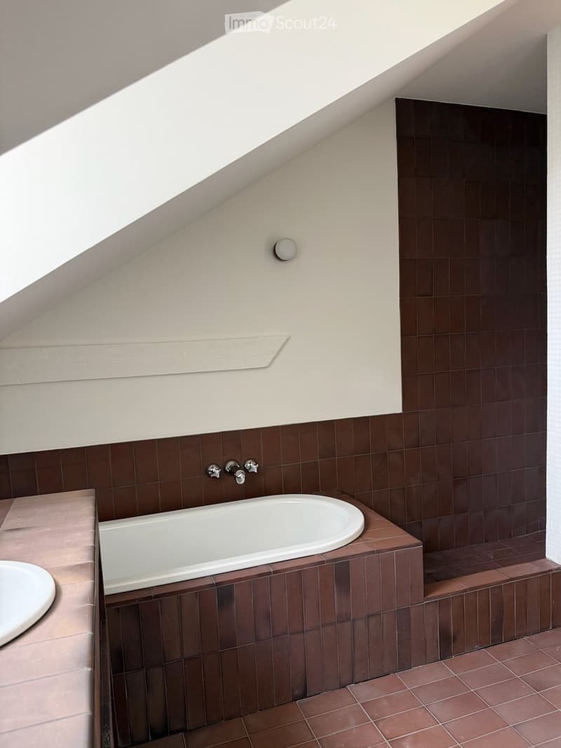 Modern bathroom with a bathtub, faucet, and sink, featuring brown tiles and a sloped ceiling with skylight.