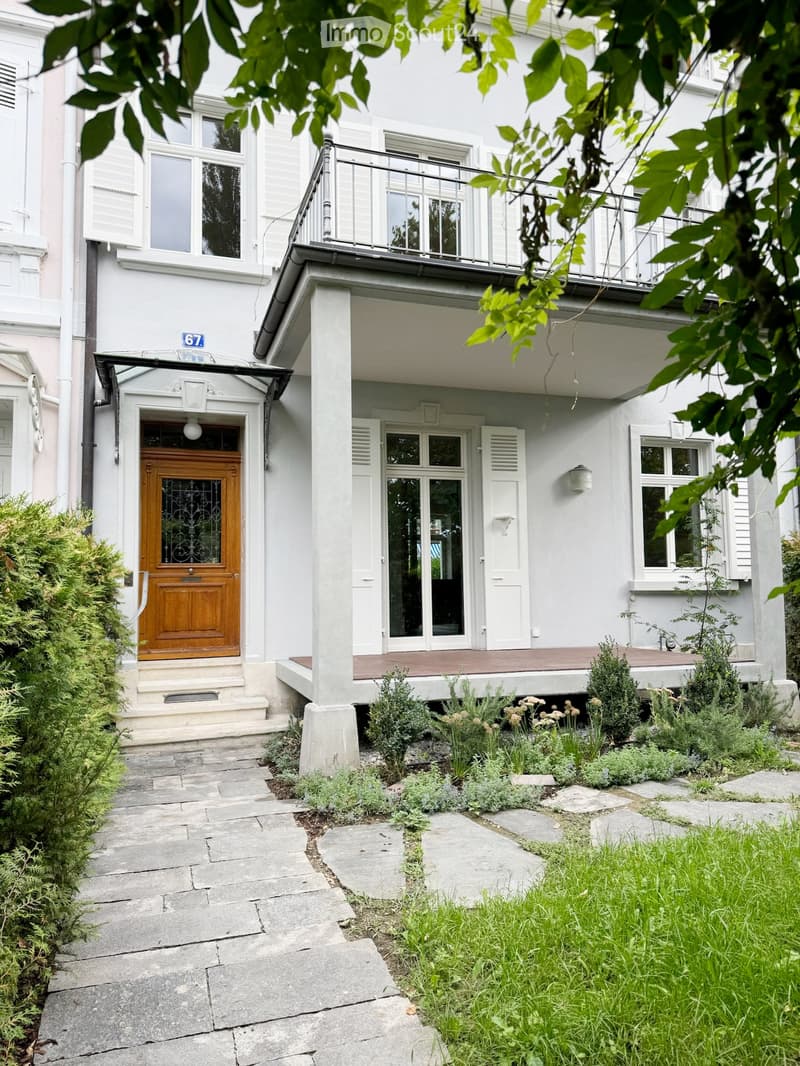 Two-story white building with a wooden front door, balcony, and small garden area with plants and shrubs.