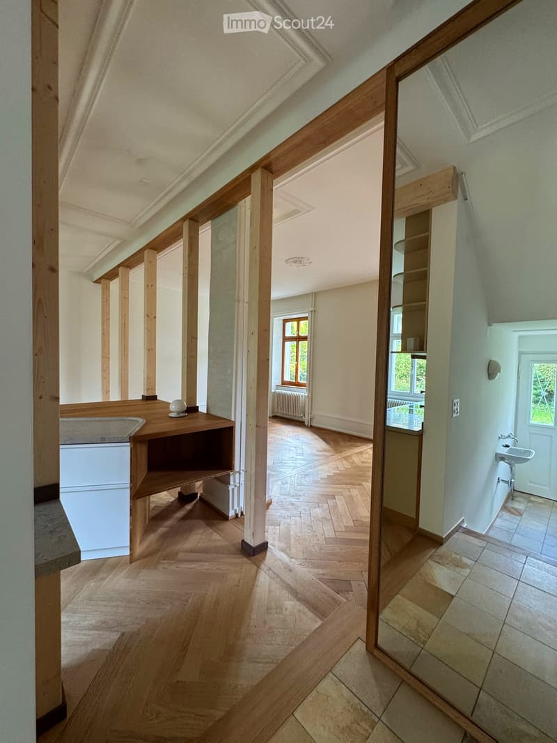 Spacious hallway with wooden flooring, wooden beams, and a small wooden shelf with a decorative item. The hallway leads to a room with a sink and a window.