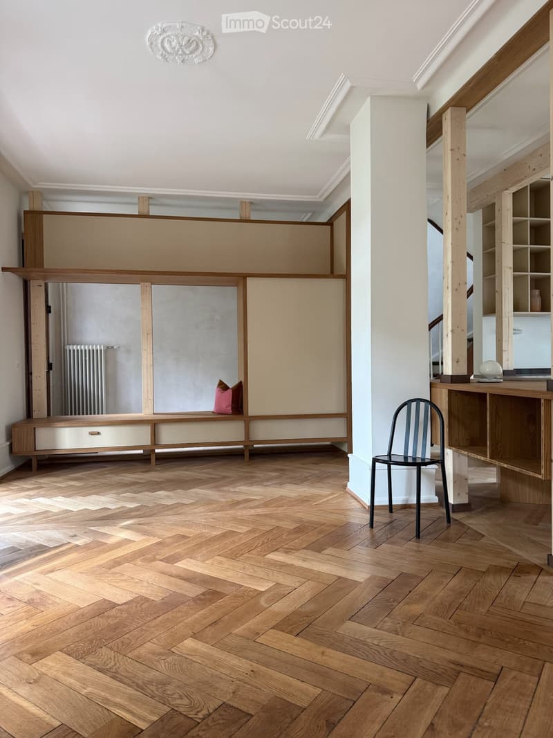 Spacious room with herringbone wooden flooring, built-in bench seating with pink cushions, a modern black chair, and a wooden shelf unit with glass compartments.