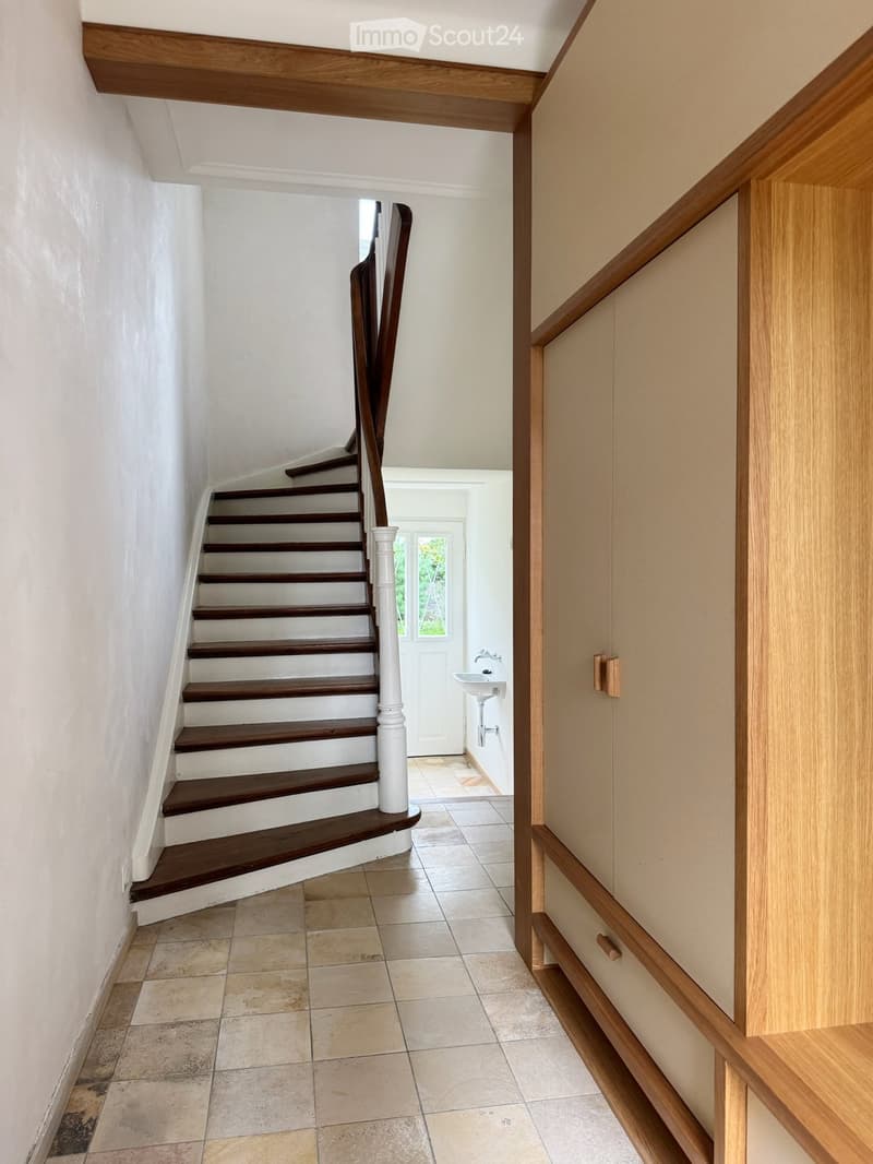 Spacious hallway with wooden staircase, white walls, tiled floor, and a small sink visible in the background.