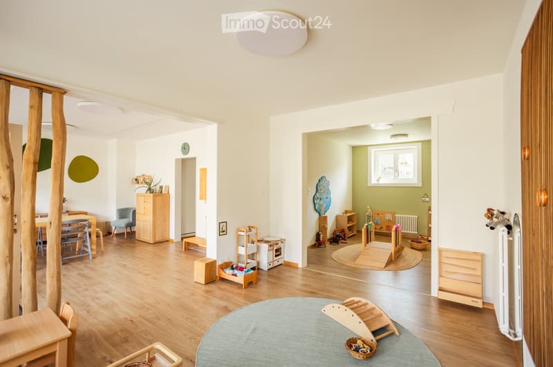 Bright, open-plan living and play area with wooden flooring, modern furniture, and various toys and play structures.