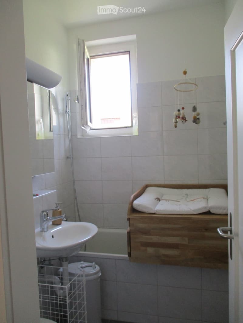 Bathroom with white tiled walls, a sink with a faucet, a mirror, a toilet, a bathtub, a wooden shelf with a cushioned surface, and a hanging toy mobile.