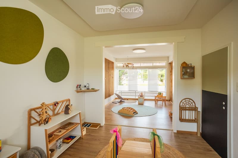Bright, open-plan room with wooden flooring, large windows, and modern decor. Features include a shelf with toys and books, a small table with chairs, a round rug, and a cabinet with glass doors.