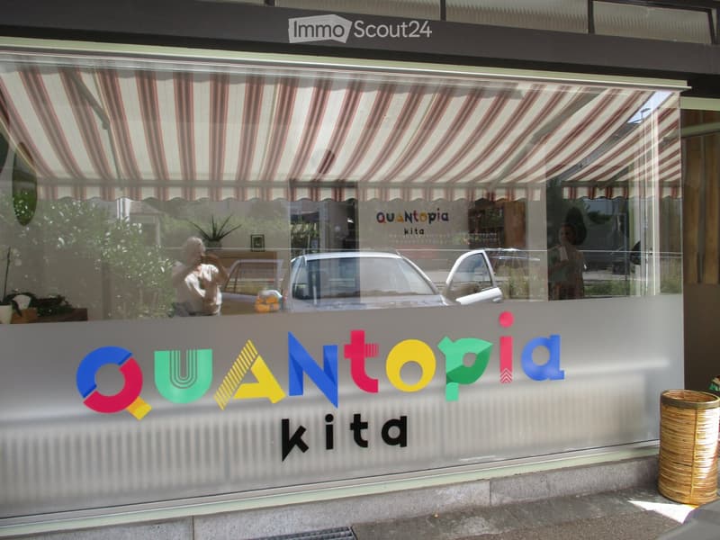 Storefront with large glass windows and colorful signage reading "QUANTOPiA kita". Visible interior includes a potted plant, a person taking a photo, and a car reflected in the glass.