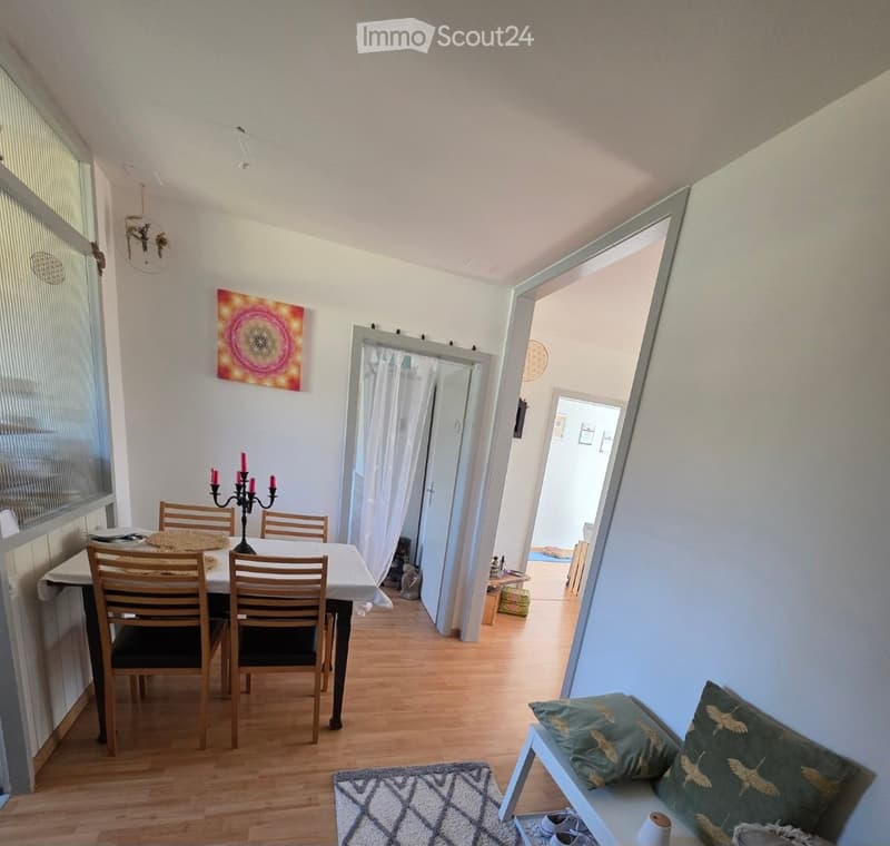 Dining area with a wooden dining table, chairs, a candle holder, a bench with pillows, and a rug on the floor. The room has light-colored walls, wooden flooring, and large windows with blinds.