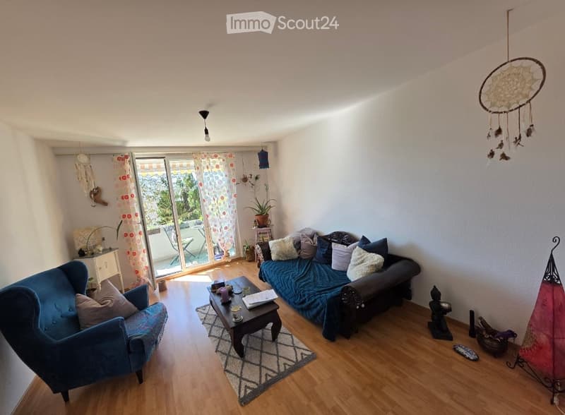 Living room with wooden flooring, large window with balcony access, sofa, armchair, coffee table, and decorative items including a dreamcatcher and plants.