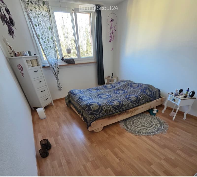 Cozy bedroom with wooden flooring, a large bed with a patterned blue and black bedspread, a nightstand with various items, a white dresser with drawers, and a dreamcatcher hanging on the wall. The room features large windows with sheer curtains and a roun