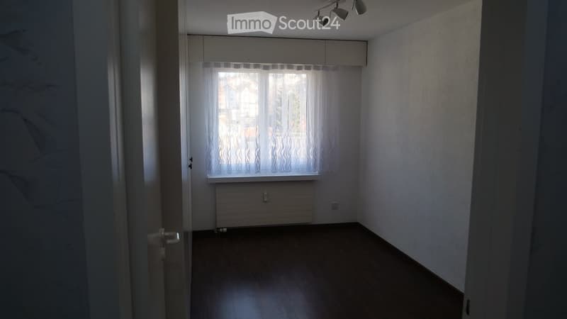 Empty room with wooden flooring, white walls, large window with white curtains, radiator, and ceiling lights.