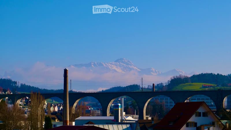 A scenic view of a town with a large bridge spanning a valley, surrounded by houses and trees, with snow-capped mountains in the background.