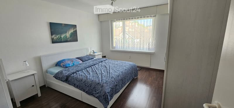 Cozy bedroom with a white bed, blue bedding, nightstand with lamp, and a picture frame on the wall. Wooden flooring and large window with blinds.