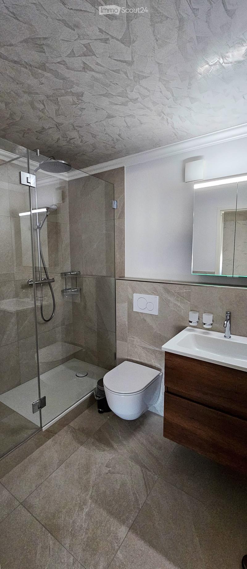 Modern bathroom with marble walls and floor, featuring a glass-enclosed shower, toilet, sink with wooden cabinet, and a large mirror above the sink.