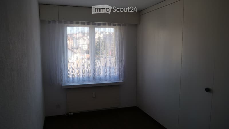 Empty room with white walls, a window with sheer curtains, a radiator, and a door.