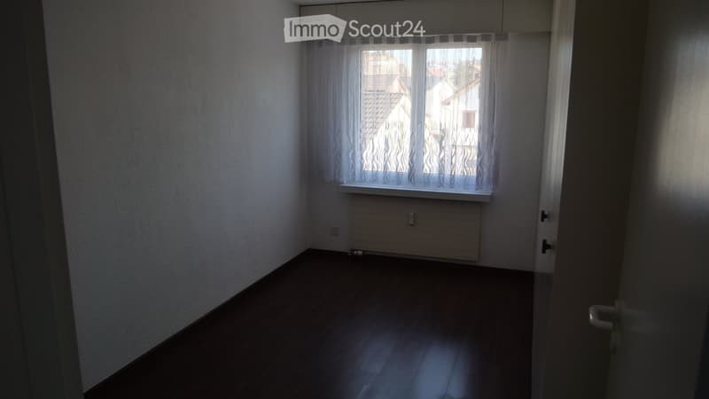 Empty room with wooden flooring, white walls, a window with sheer curtains, and a radiator below the window.