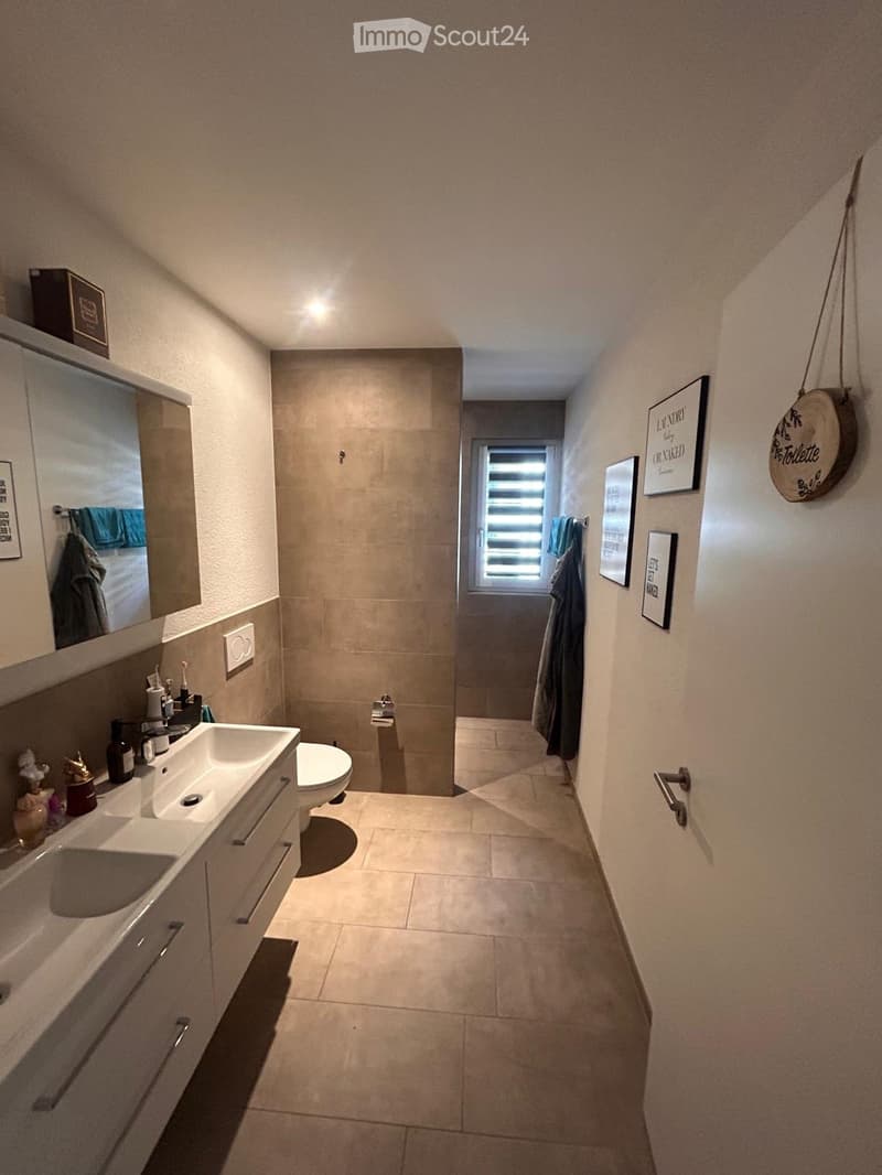 Modern bathroom with a double sink, toilet, mirror, and framed pictures on the walls. The space features beige tiles and a window with blinds.
