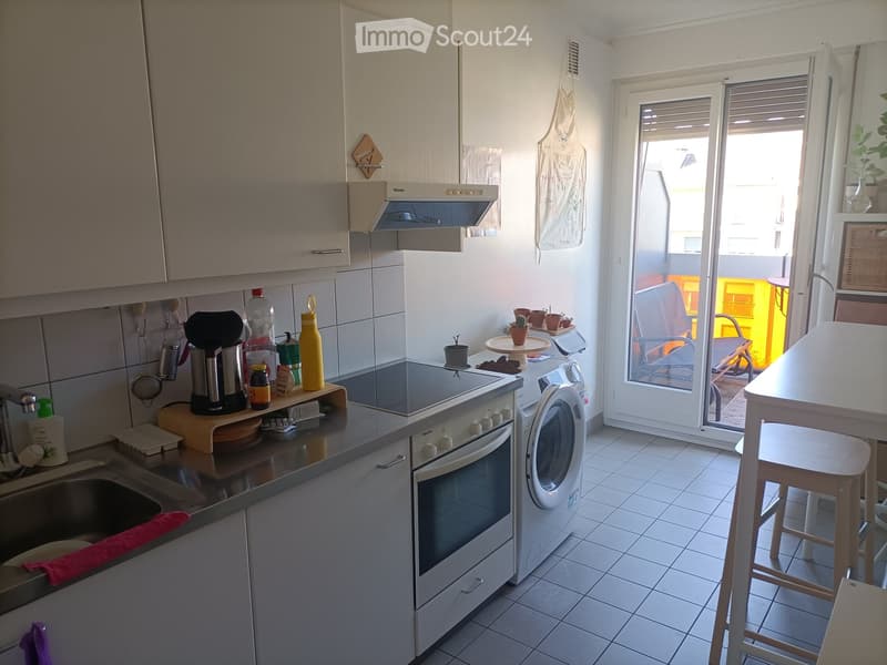 Modern kitchen with white cabinets, stainless steel appliances, and a small dining area with a table and stools. A sliding glass door leads to a balcony with outdoor furniture.