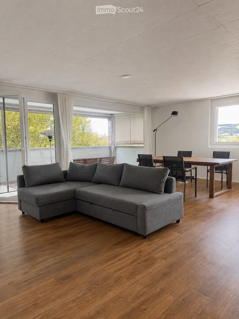 Modern living and dining area with a grey sectional sofa, wooden dining table, and chairs, large windows with curtains, and a view of greenery outside.
