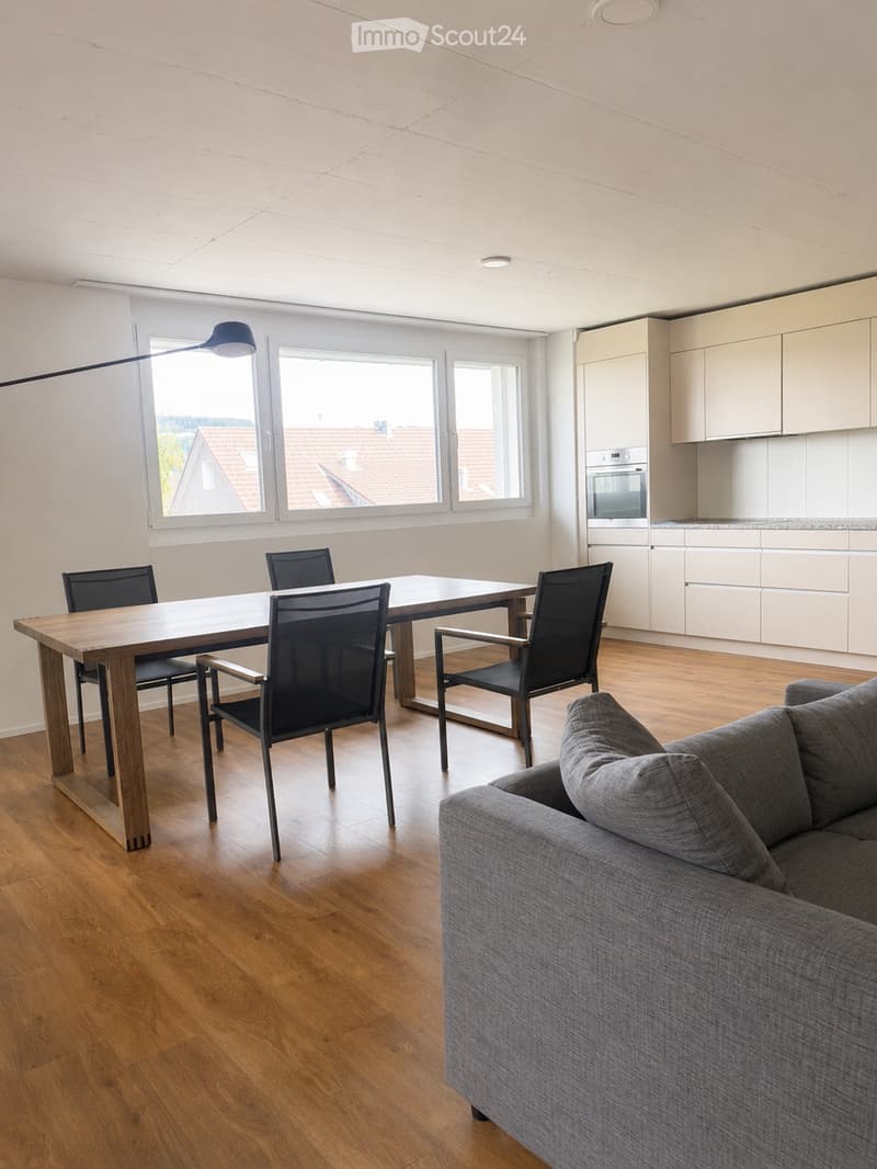Open-plan living and dining area with a grey sofa, wooden dining table, four chairs, and a modern kitchen with white cabinets and an oven.