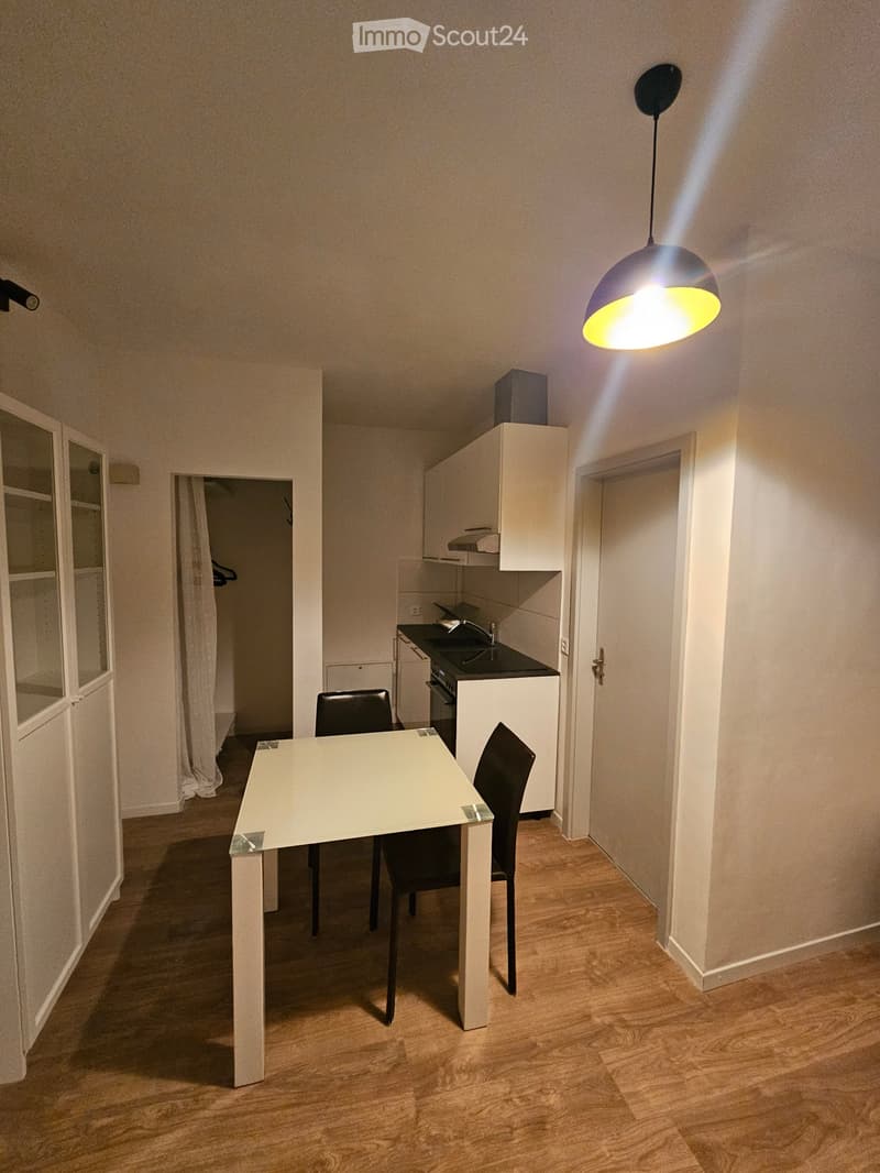 A modern, well-lit room with a white dining table and two black chairs, a small kitchen area with cabinets, an oven, and a sink, and a hanging light fixture.
