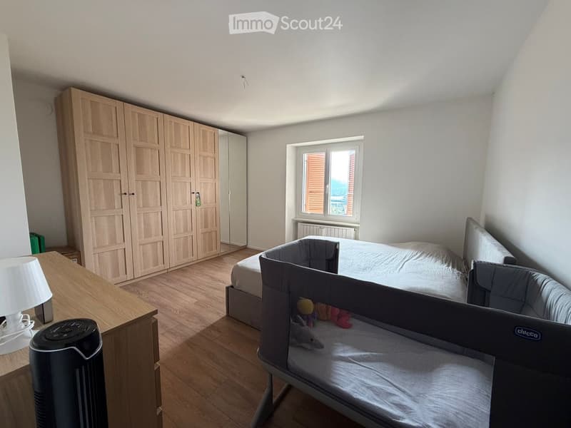 Bright bedroom with wooden floor, large wardrobe, bed with bedding, baby crib, and a small table with a lamp.