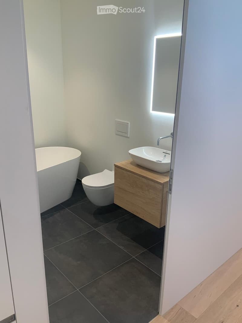 Modern bathroom with dark tiled floor, white bathtub, toilet, and sink on a wooden cabinet.