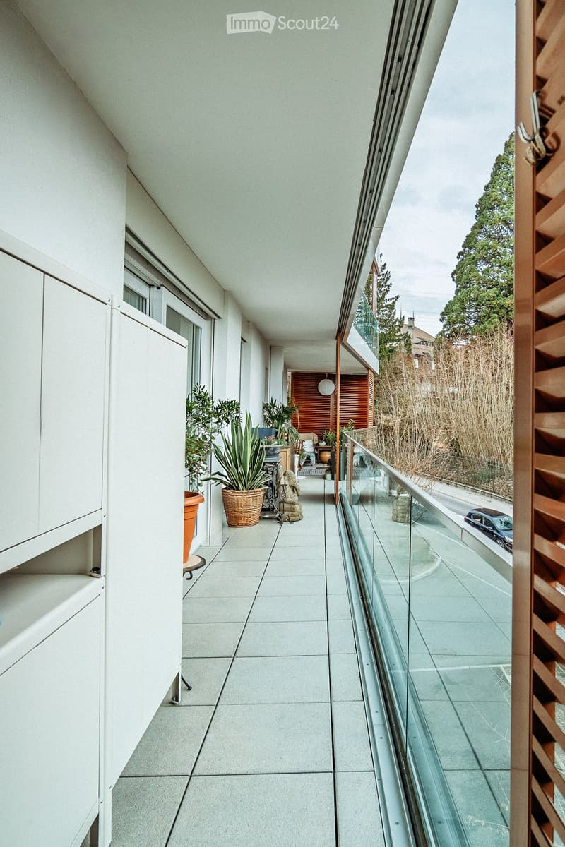 Modern balcony with glass railing, tiled floor, and potted plants, overlooking a street with a parked car.