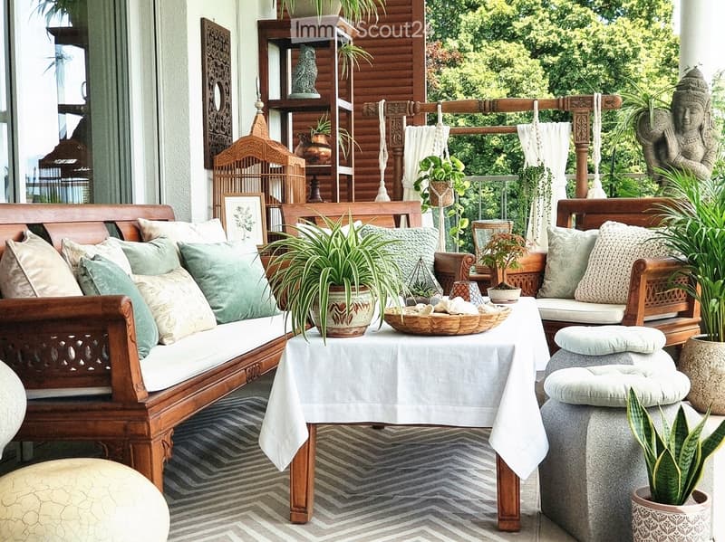 A well-decorated balcony with wooden furniture, cushions, potted plants, and a small table with a white tablecloth.