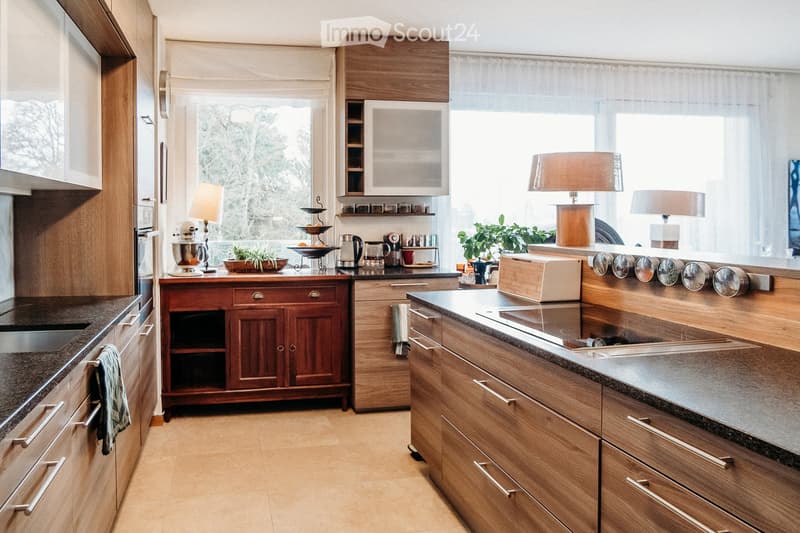 Modern kitchen with wooden cabinets, stainless steel appliances, granite countertops, and a large window with sheer curtains. Includes a sink, oven, microwave, refrigerator, blender, coffee maker, toaster, and decorative items on the counter.