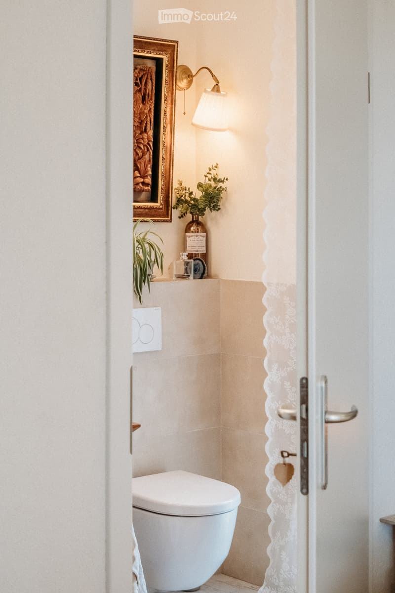 Modern bathroom with a toilet, wall-mounted lamp, framed picture, small plant, and decorative items on a shelf.