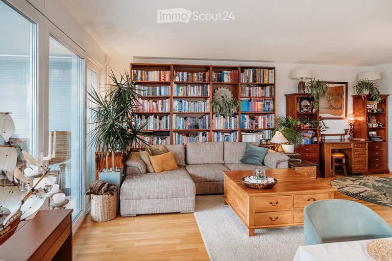 Modern living room with a grey sectional sofa, wooden coffee table, bookshelves, desk, and decorative plants.