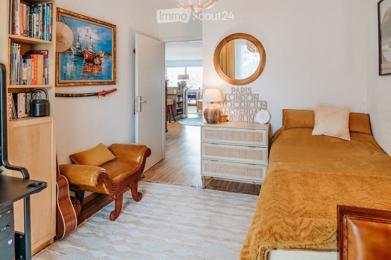Cozy bedroom with a bed, leather armchair, nightstand with lamp, bookshelf, and a round mirror on the wall. The room features warm tones and a mix of modern and vintage decor.