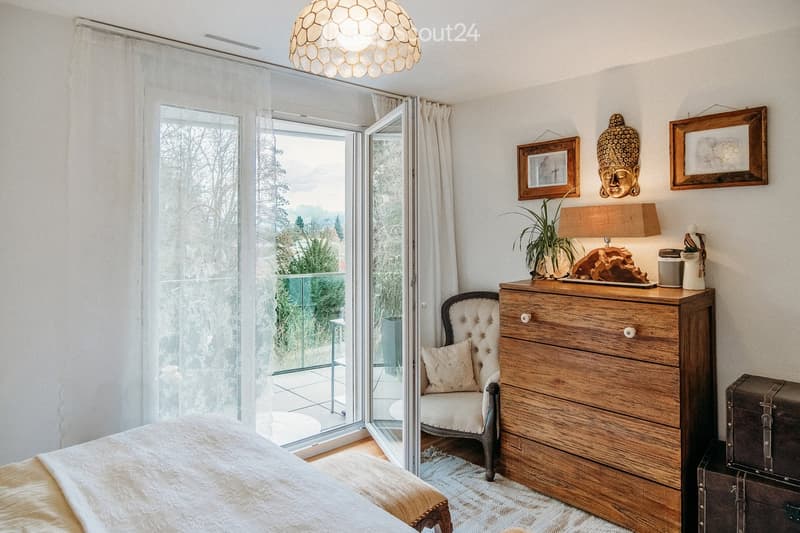 Bright bedroom with a large bed, a tufted armchair, a wooden dresser, a table lamp, and a decorative plant. Large windows open to a balcony with a view of greenery.