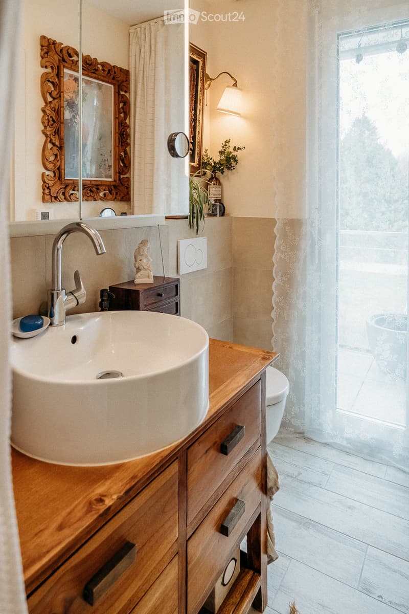 Elegant bathroom with a large wooden vanity, a modern sink, a stylish mirror, and a toilet. The space features natural light from a window with sheer curtains and decorative elements like a framed picture and a small plant.